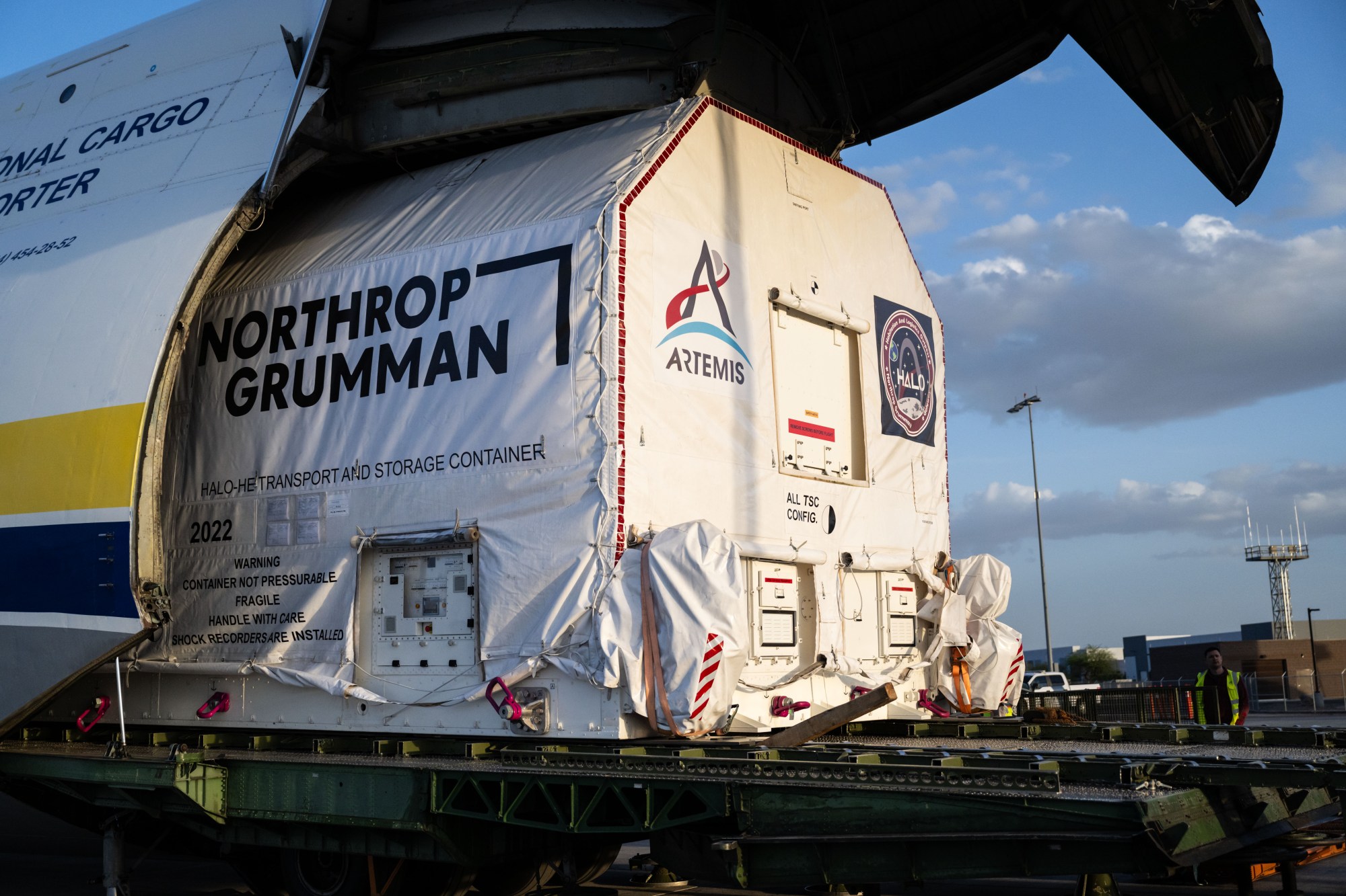 A large cargo aircraft with its nose lifted open reveals a wrapped container being offloaded. The container is marked with the logo for NASA's Artemis campaign and labeled as the HALO transport and storage container. The scene is set at an airport during late afternoon.