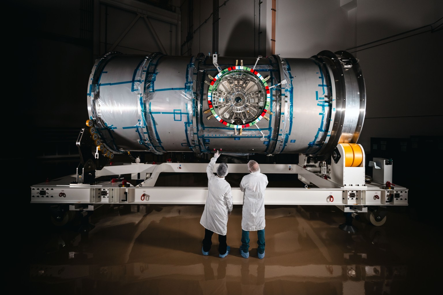 Two technicians in cleanroom attire stand in front of HALO (Habitation and Logistics Outpost) at Northrop Grumman’s facility in Gilbert, Arizona. The cylindrical metallic module, recently unboxed, rests on a white stand. One technician points toward the front of the module.