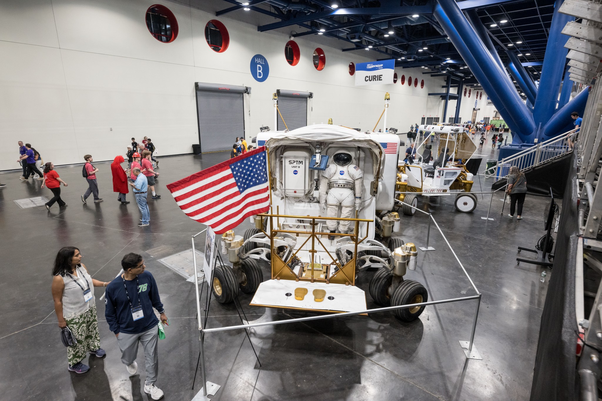 Visitors walk through an indoor exhibit featuring a large NASA space exploration vehicle with a mounted spacesuit and American flag.