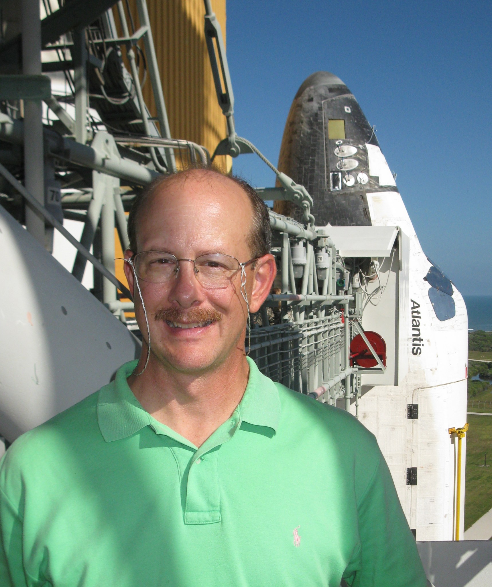 A man wearing a neon green polo smiles outside on a sunny day in front of a space shuttle.
