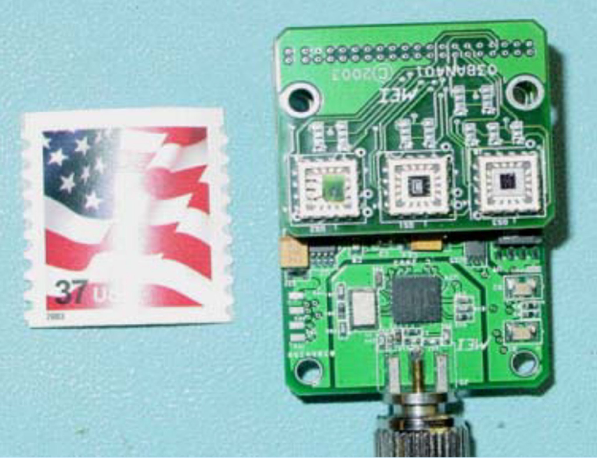 A small electronic circuit board with various components is displayed beside a U.S. flag postage stamp to compare size. The background is a light blue surface.