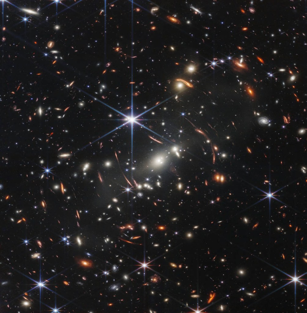 In this deep field image, many galaxies are visible against a black backdrop. Some appear as only pinpricks of light. Near the center of the image, one object shines brightly. Some objects appear warped and have a redder color than other objects visible in the image.