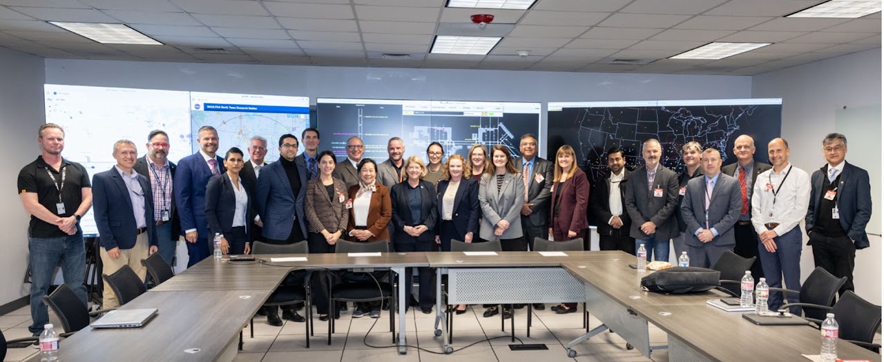 Group photo of Pam Melroy with the ATM-X team standing in front of 3 large monitors and behind a U-Shaped table.