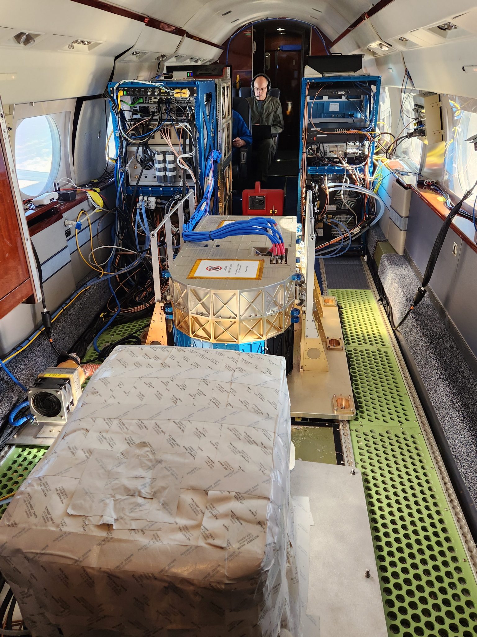 A person can be seen standing in the rear of the photo looking out at large oblong and box shaped instruments on the floorboard of a specially modified aircraft.