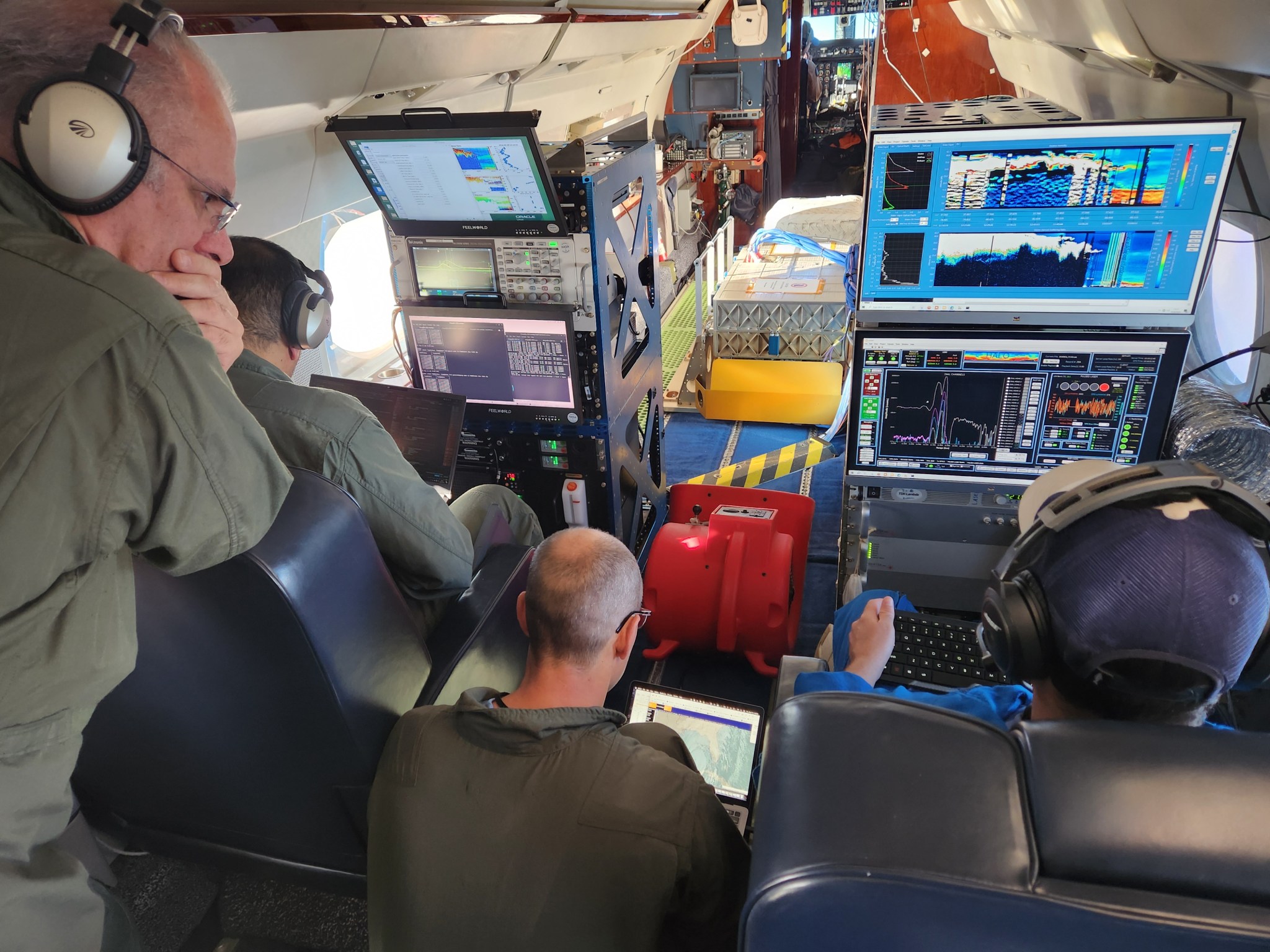 A man wearing ear protection headphones can be seen standing near two men sitting as they all look at data screens while flying science mission flights for AWP.