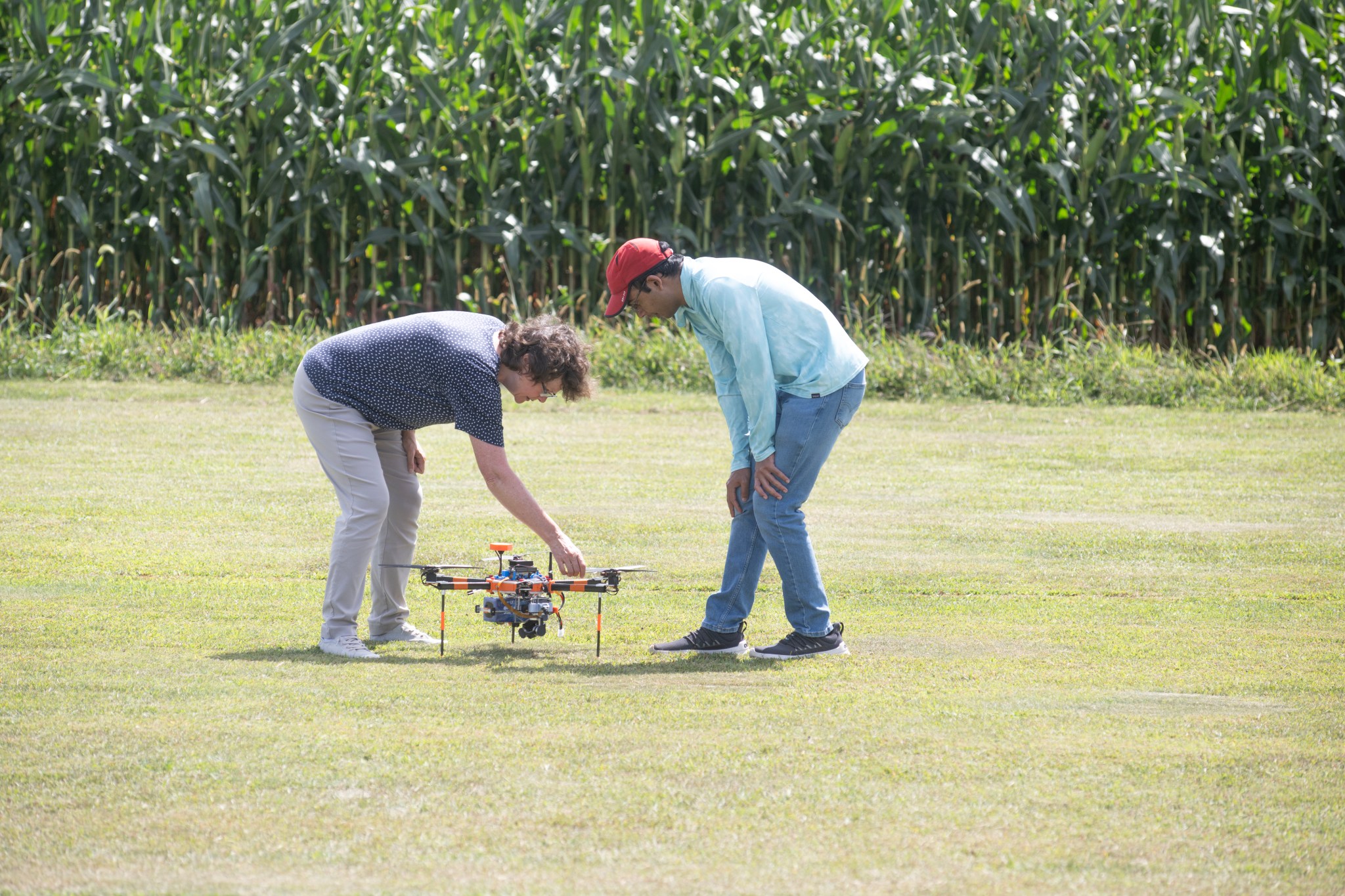 In the foreground two men bend down and look at a drone on the ground. The drone is sitting on grass. The man on the left side reaches down to touch the drone. The drone is orange and black. The man on the left side is wearing light grey pants and a dark grey shirt. He has brown, curly hair and is wearing white tennis shoes. The man on the right side wears black tennis shoes, a red baseball cap, jeans and a light blue shirt. In the background there is a line of corn plants.