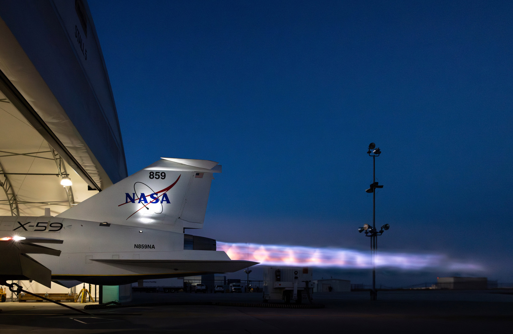 A NASA X-59 aircraft sits on the tarmac at dusk with its engine running, producing a bright, fiery exhaust plume trailing into the night sky. The aircraft is partly inside a hangar.