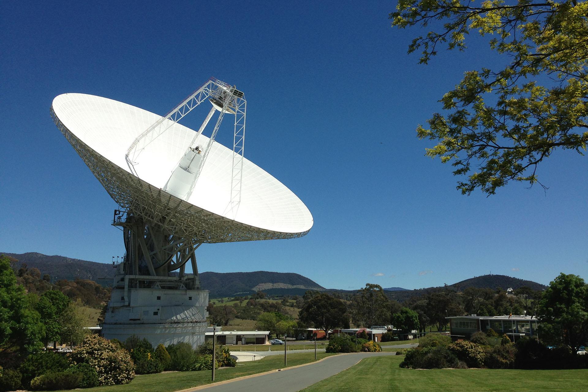 A massive white radio antenna's dish points up and to the right in this photo. There is grass and many trees all around it. Hilly terrain is in the background, rising up to meet the blue sky.