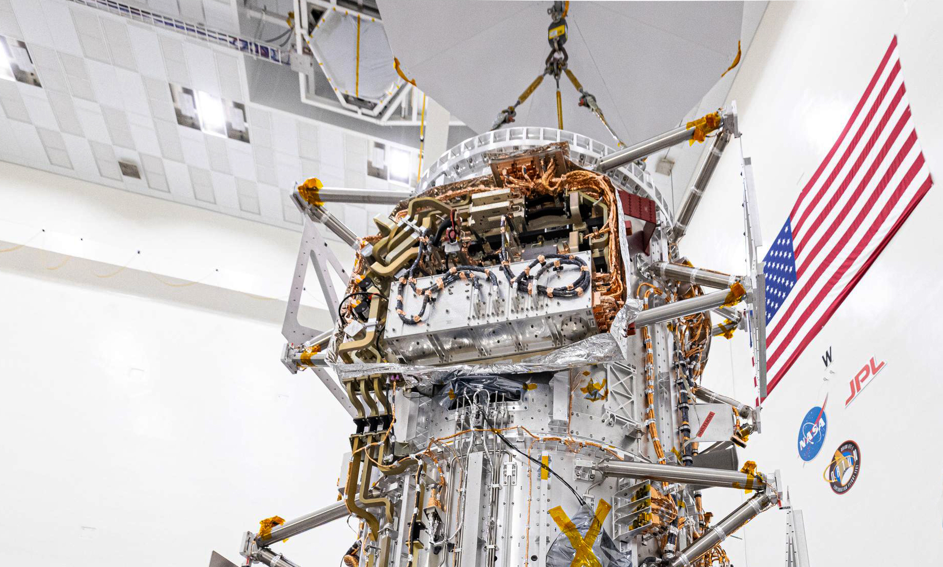 In a clean room, Europa Clipper is examined by engineers, the photo is focusing on a box full of wires toward the top of the spacecraft
