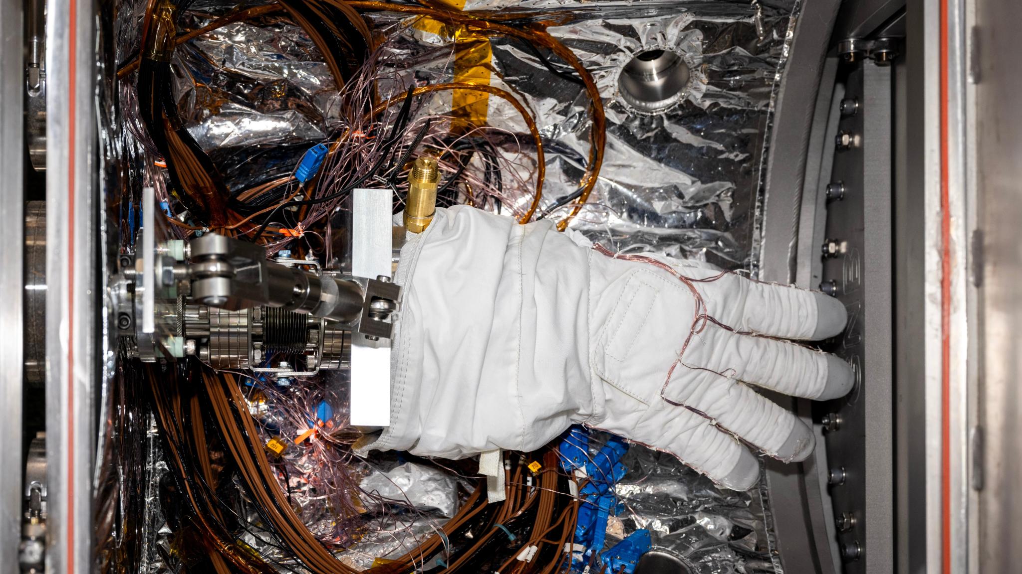 A bulky white spacesuit glove with gray fingertips sits inside a small chamber lined with silvery material. Multiple wires wrap around the fingers of the glove and loop around the floor of the chamber beneath the glove.