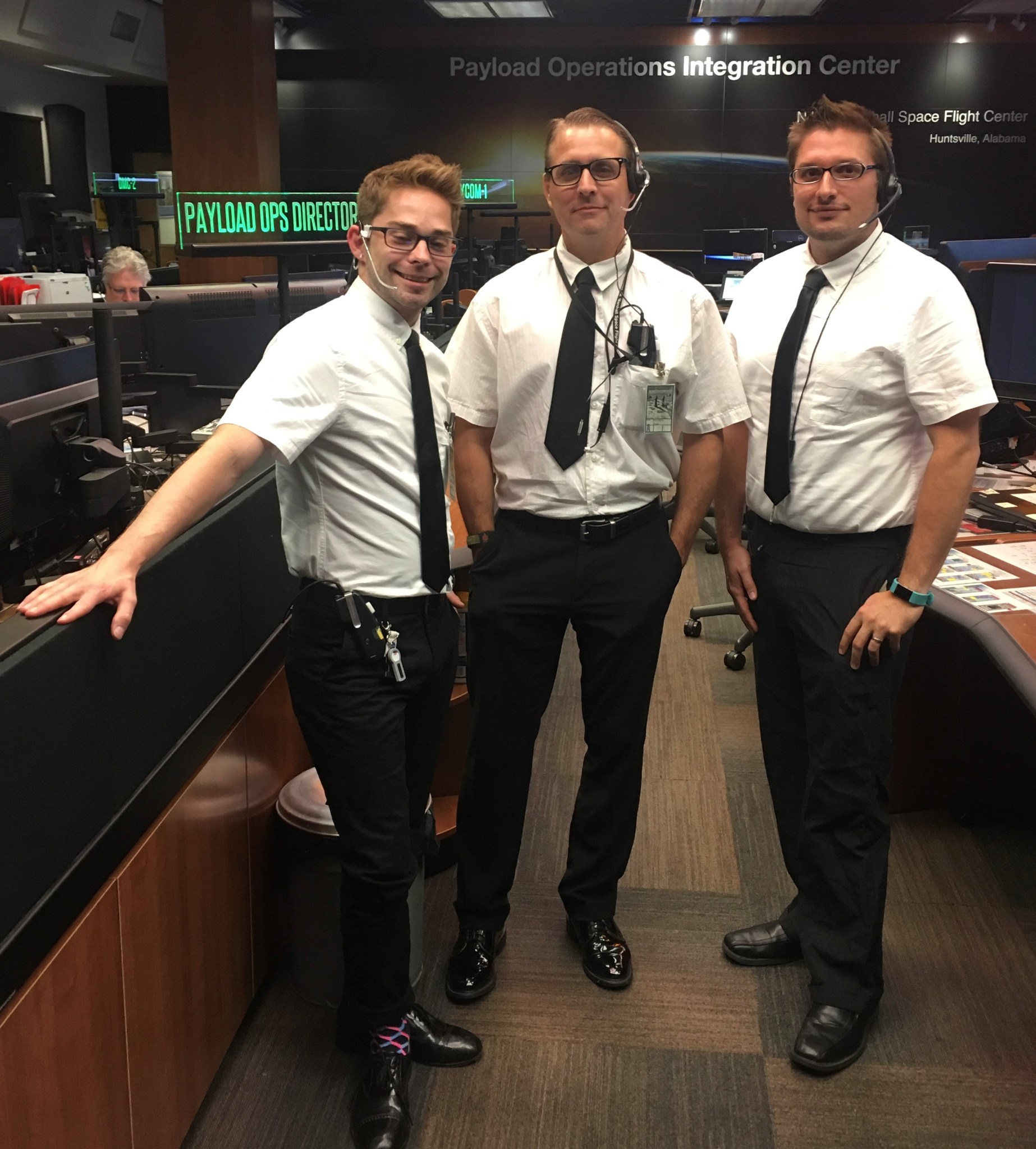 Three men wearing white button downs and black tie and pants stand inside of a control room.