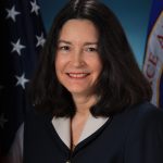 Image shows woman in black suit jacket posing for a photo with American flag in the background.
