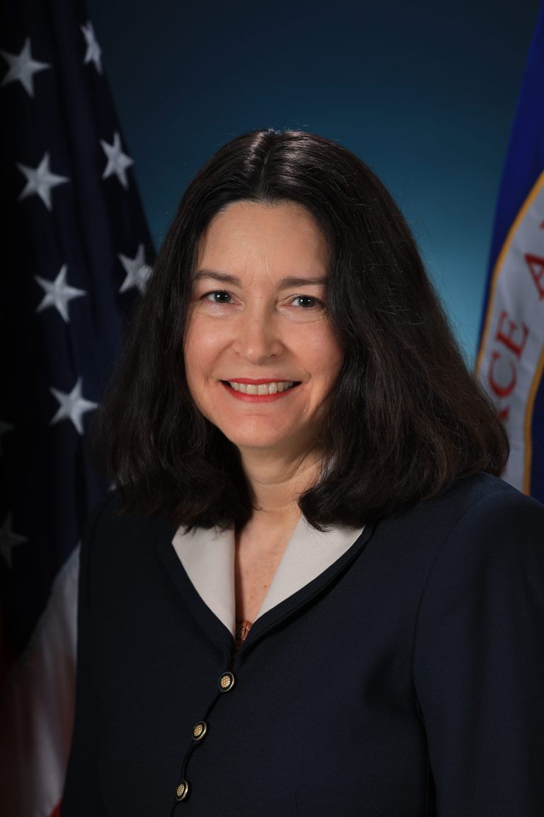Image shows woman in black suit jacket posing for a photo with American flag in the background.