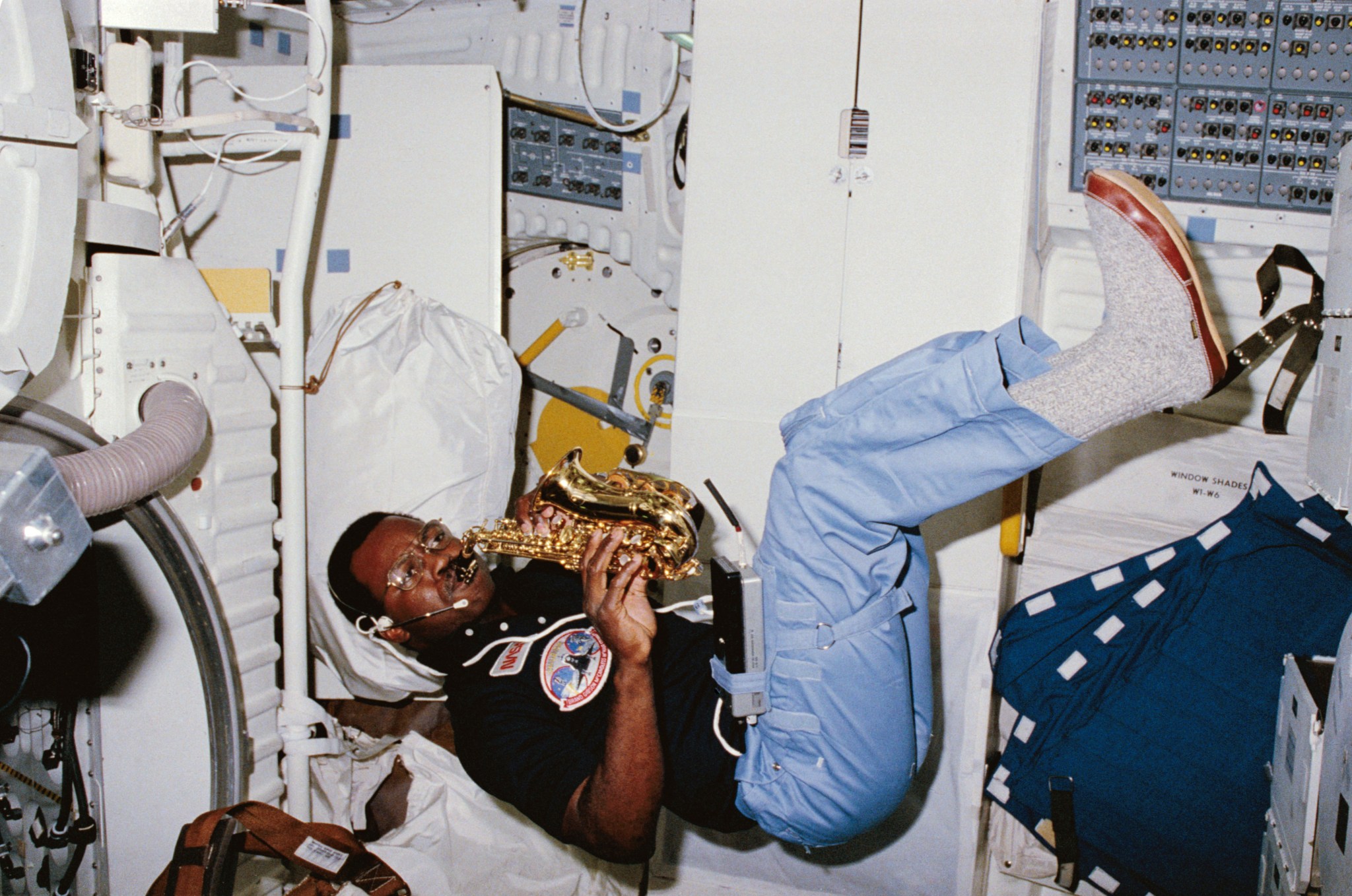 A Black man plays a saxophone while floating. He wears a black short sleeve shirt with patches on it, blue pants, and gray shoes. He is inside the space shuttle Challenger.
