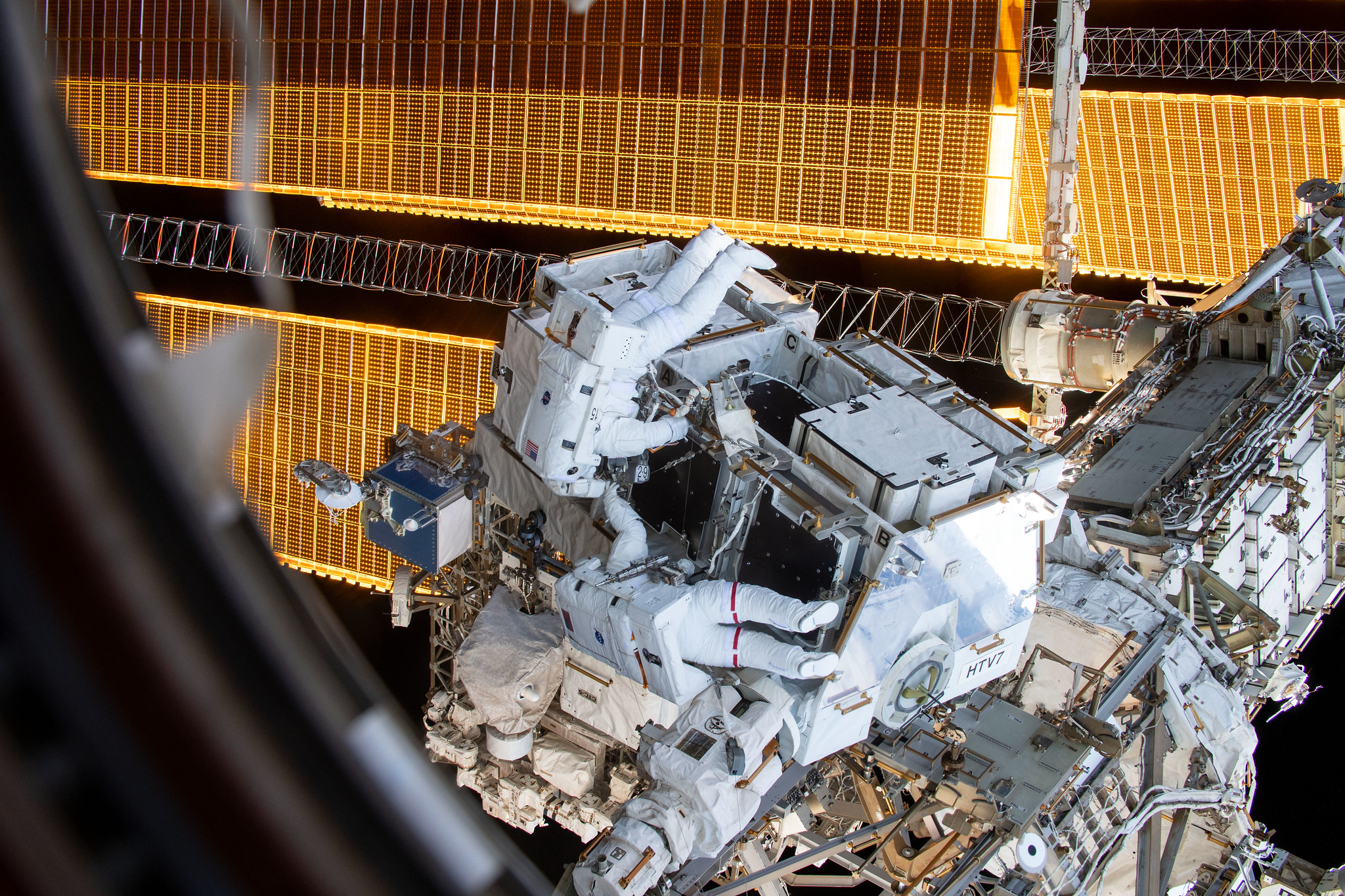 Two astronauts are seen working on the outside of the International Space Station through a window.