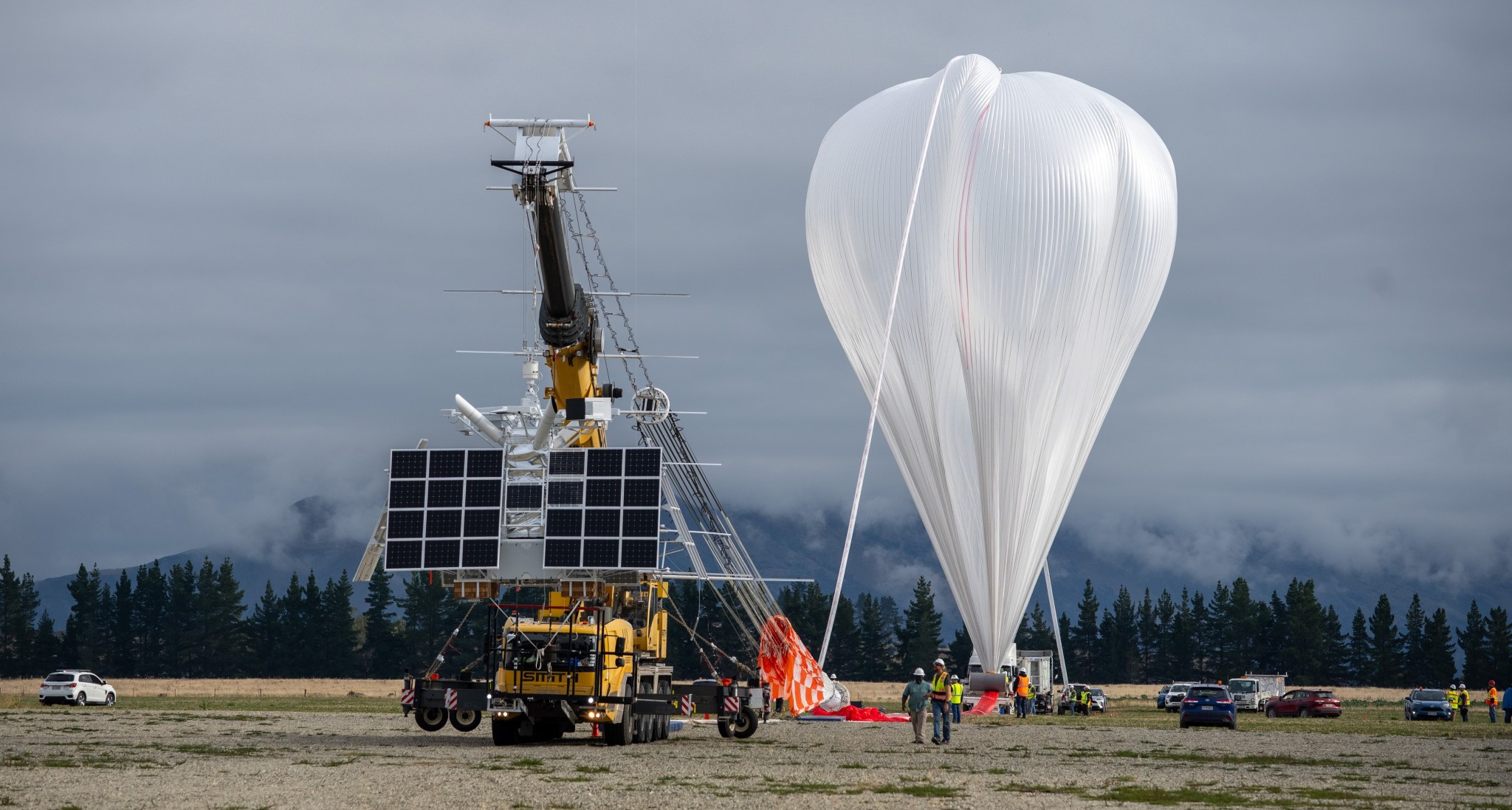 A large metal scientific instrument with black solar panels is suspended off the ground by a yellow crane. A white trail of material is on the ground and attaches to a partially inflated white balloon in the background.