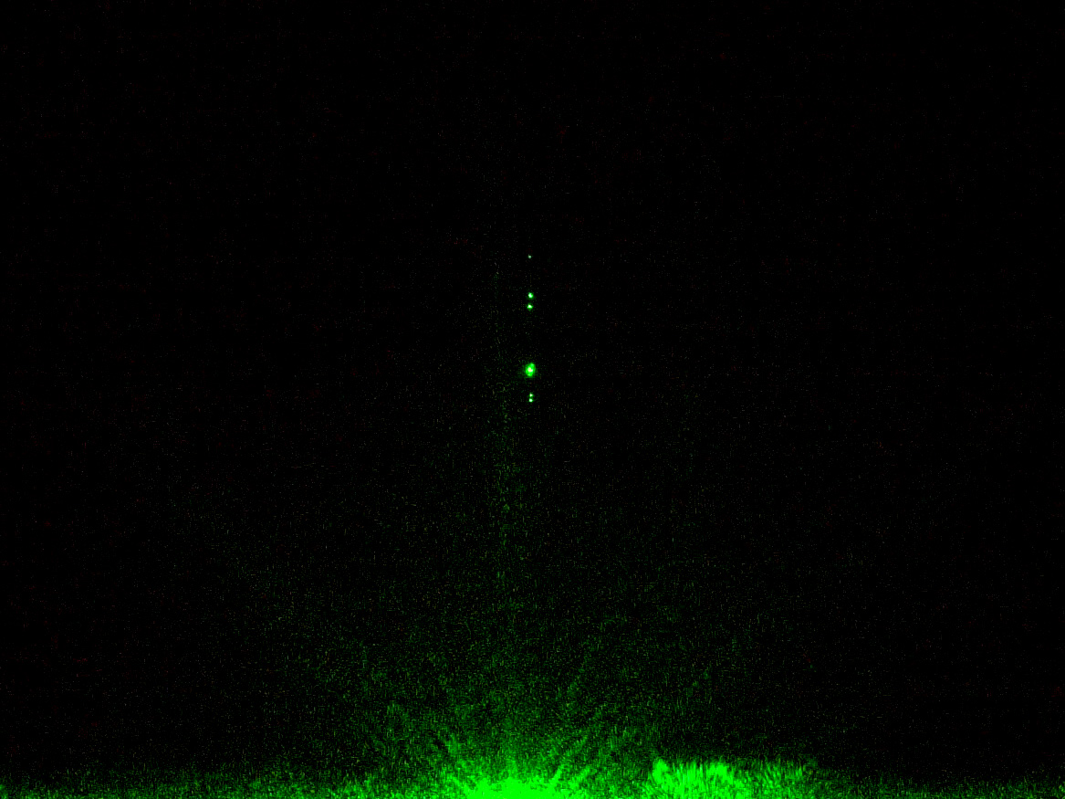 Small spheres seen as points of light above a glass plate with a mirror underneath, illuminated in green laser light with a black background.