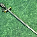 A long, metallic pipe-like device with a cylindrical component and yellow caps on one end, lying diagonally on a green marbled surface.