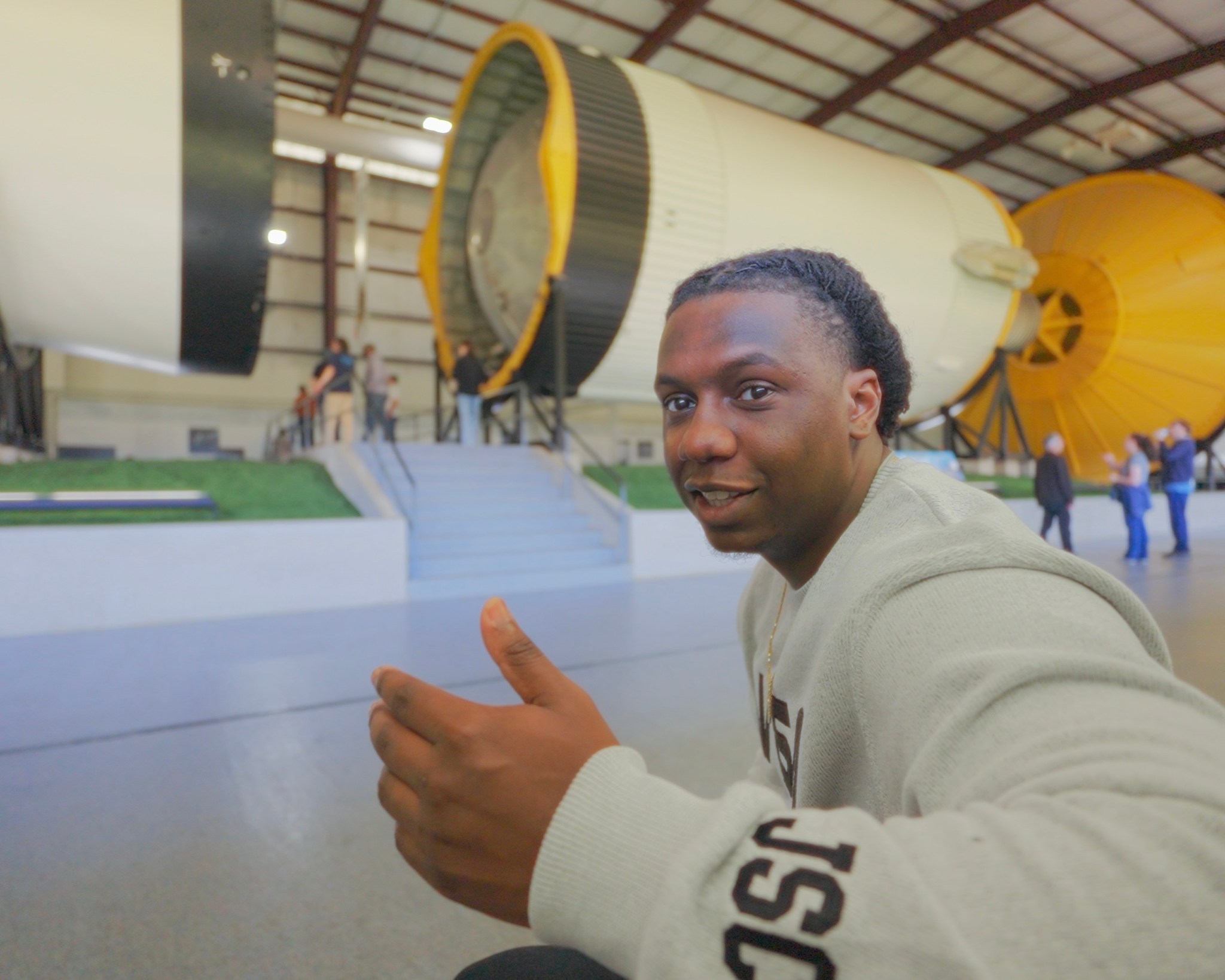 A smiling man in a light gray NASA sweatshirt gestures with a thumbs-up while seated in front of a massive Saturn V rocket display.