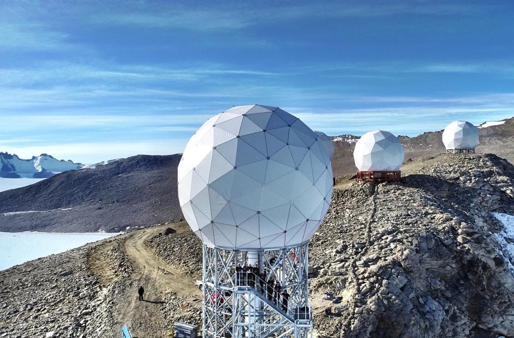 Three white, geodesic radomes stand on elevated platforms at the Troll Satellite Station in Antarctica. The radomes protect antennas positioned along a rocky, snowy ridge. The surrounding landscape features rugged terrain, distant mountains, and a bright sky, while a line of smiling KSAT employees line up on the staircase leading up to the antenna’s radome. The antenna in the foreground, TR9, was upgraded to support the Ka-band signal and a cloud storage system in preparation for SPHEREx.