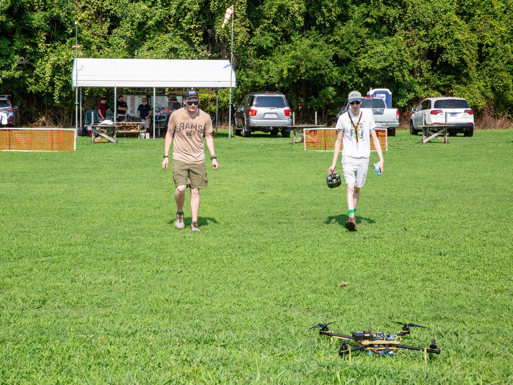 A drone sits on the ground in the foreground of the image. The ground is bright green grass. Two men walk toward the drone, the man on the right side carrying a black controller. The man on the right side is wearing a white t-shirt and white shorts and a blue and grey baseball cap as well as brown boots and green socks. He is wearing a black and yellow lanyard and carrying a blue piece of equipment in his left hand. The other man is wearing a brown t-shirt and brown shorts. He is wearing a black watch on one hand and a brown armband on the other. He is wearing a black baseball cap and he is wearing brown boots. Both men are wearing black sunglasses. In the background there are three cars in view, a windsock on a pole and a white tent with others standing around a picnic table with various pieces of equipment. Behind this is a tall, green tree line. There are also two fences made of white pipes and orange plastic barriers in the background blocking the grass area from the parking area.