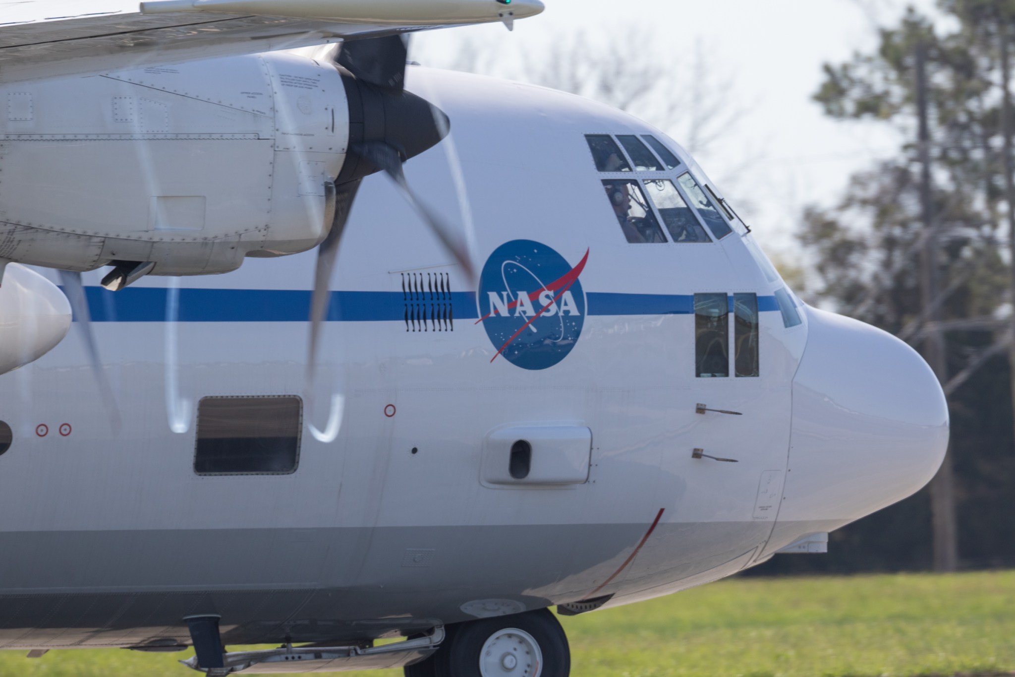 A large C-130 aircraft with a blue stripe and NASA logo on the side sits ready to take off. Large propellors are spinning and a pilot is visible from the front window.