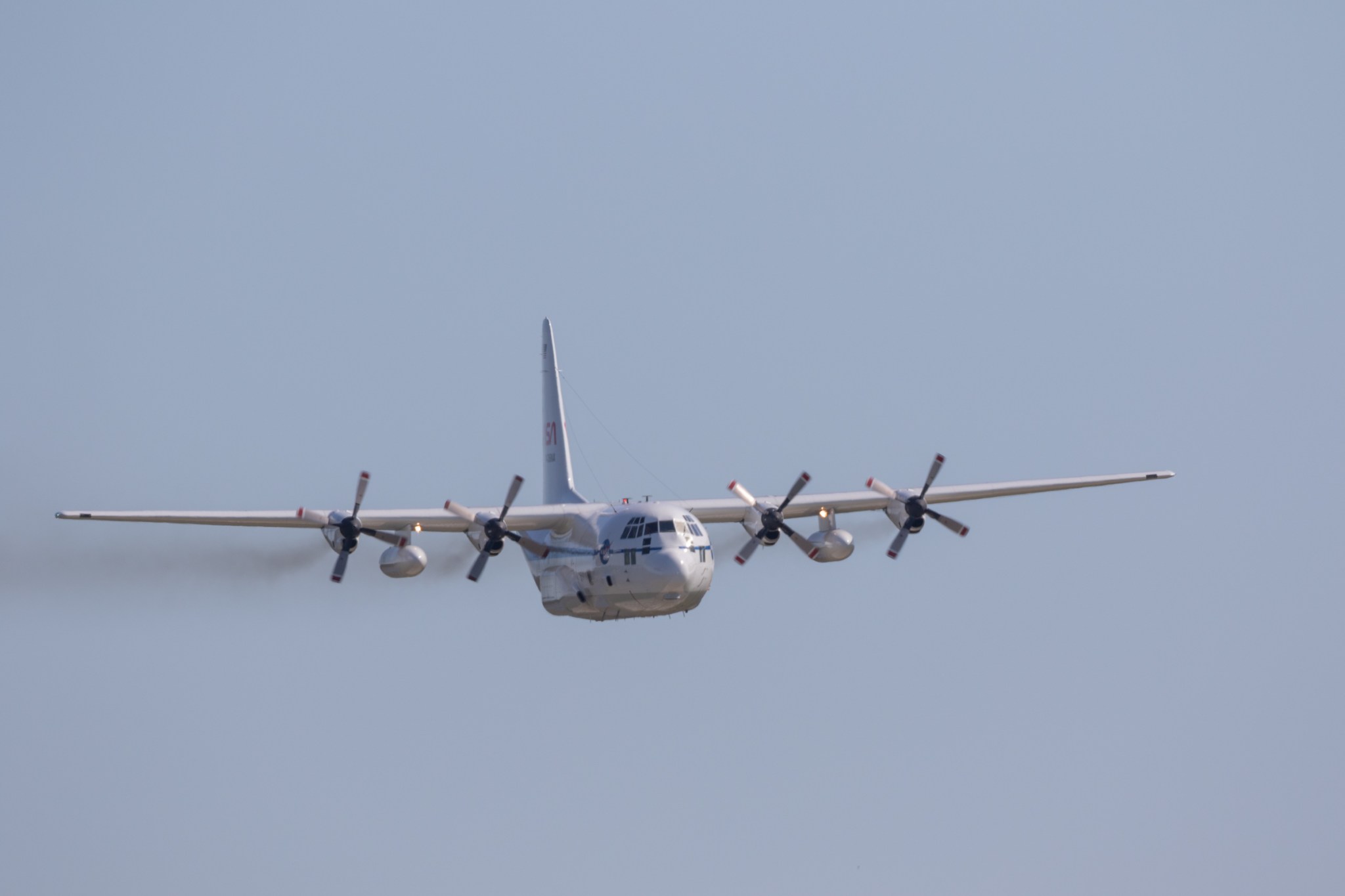The large C-130 aircraft is flying. The propellors spin against the blue sky.