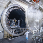 The thermal vacuum chamber door that is 20 feet tall is open. A person looking very tiny in comparison, stands in the doorway.