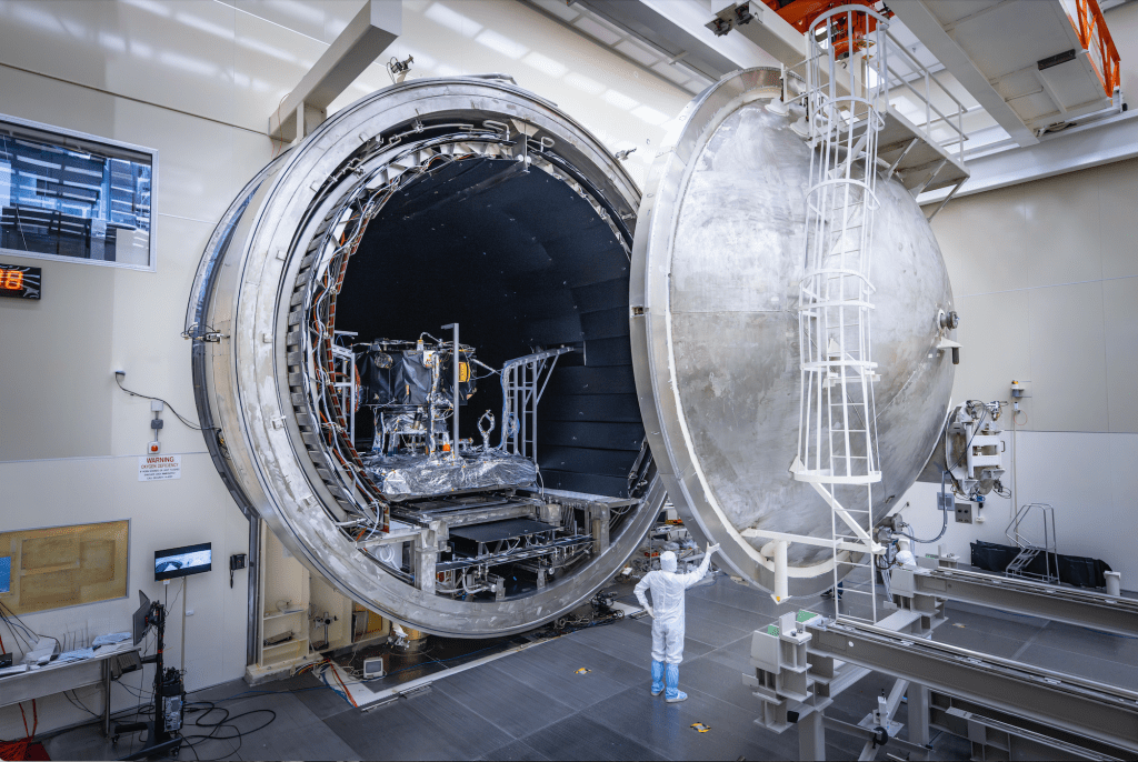 The thermal vacuum chamber door that is 20 feet tall is open. A person looking very tiny in comparison, stands in the doorway.
