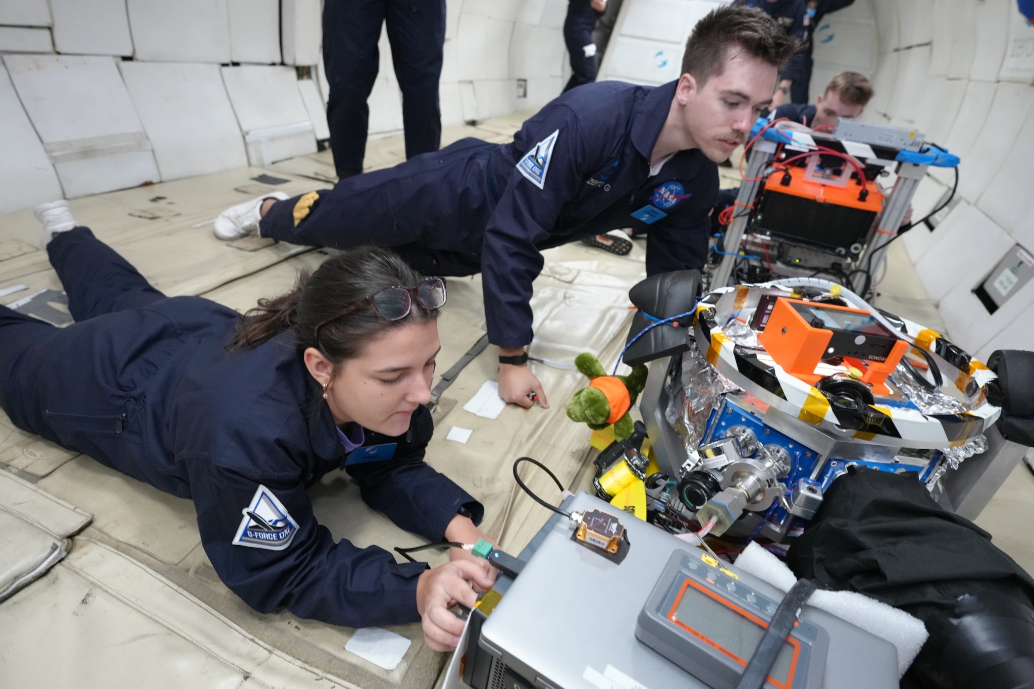 Two researchers lie on the floor of a parabolic flight making adjustments to their experiment.