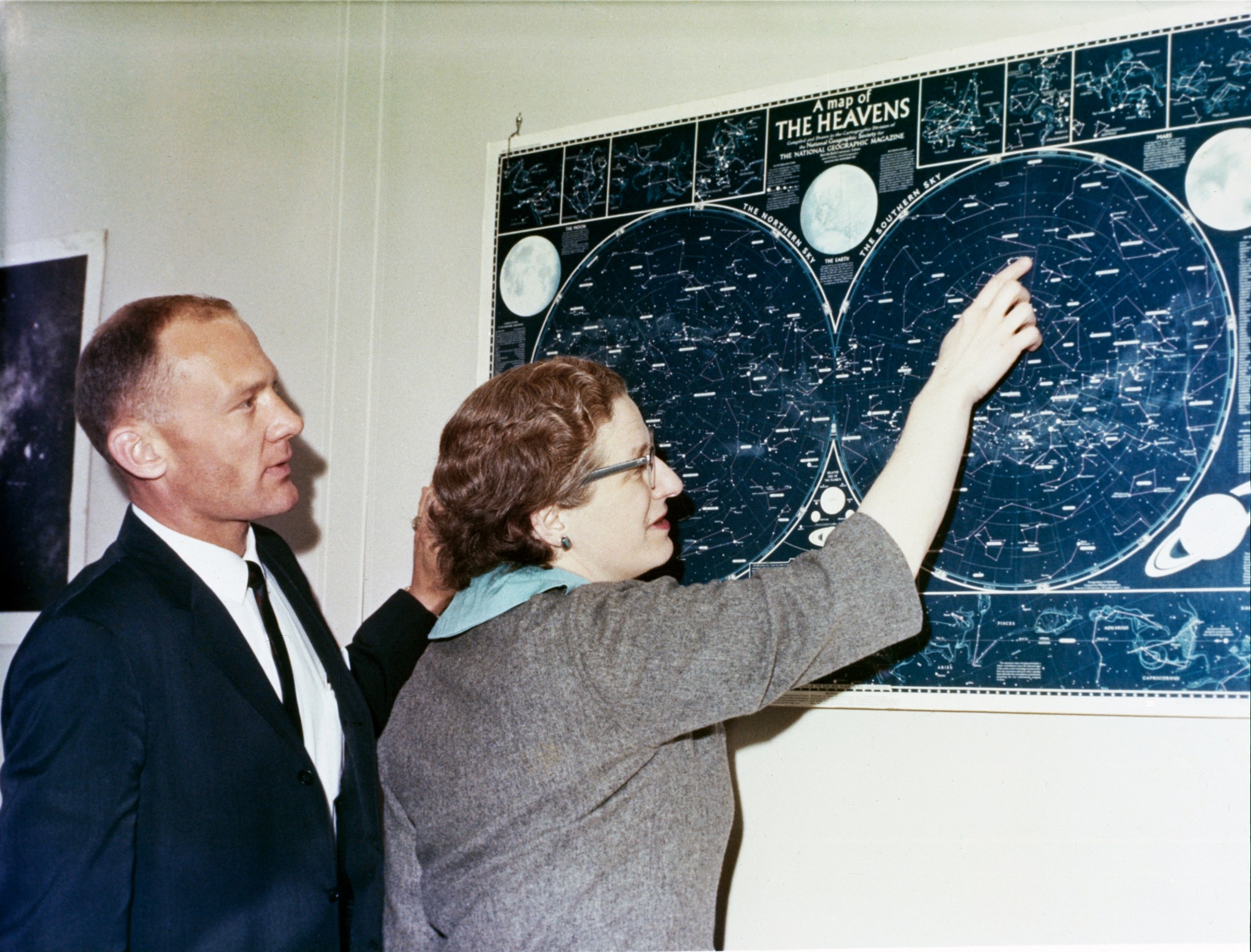 Two people - astronaut Buzz Aldrin and Dr. Nancy Grace Roman - look at a map of celestial objects hanging on a wall in front of them. The photos is taken from the side, so their profiles are visible. Aldrin wears a dark suit and tie, while Roman wears a gray short sleeved jacket. She is pointing at a constellation on the blue and white map.