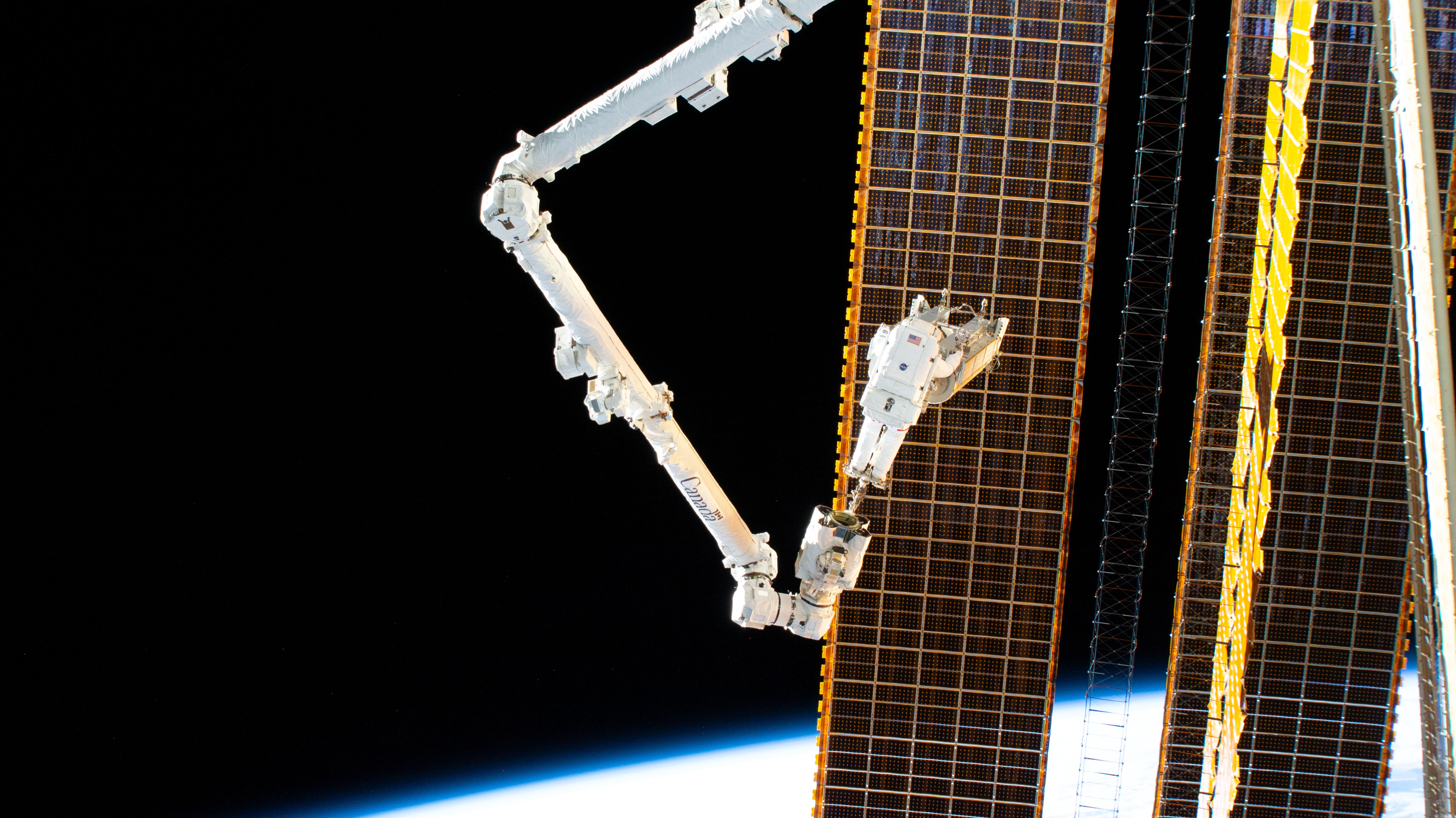 NASA astronaut Woody Hoburg is pictured during a spacewalk riding the Canadarm2 robotic arm and carrying a roll-out solar array to its installation...