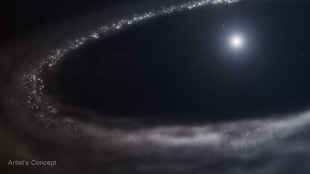 An illustration of Sun-like star HD 181327 and its surrounding debris disk. The star is at top right. It is surrounded by a far larger debris disk that forms an incomplete elliptical path and is cut off at right. There’s a huge cavity between the star and the disk. The debris disk is shown in shades of light gray. Toward the top and left, there are finer, more discrete points in a range of sizes. The disk appears hazier and smokier at the bottom. The star is bright white at center, with a hazy blue region around it. The background of space is black. The label Artist's Concept appears at lower left.