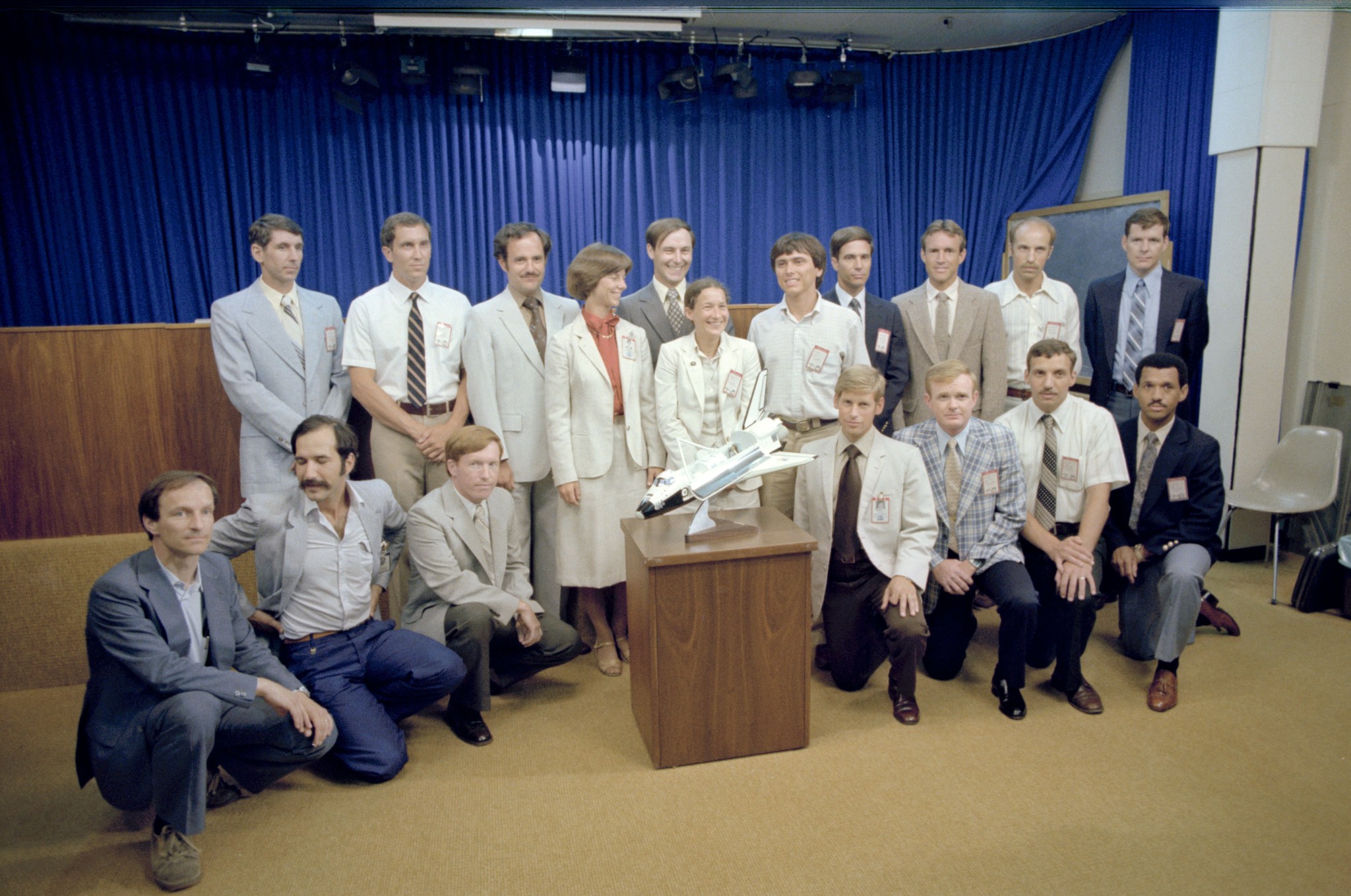 A group of people dressed in suits pose for a picture behind a model of a spacecraft. The group is organized into two rows; the first row kneels while the back row stands.