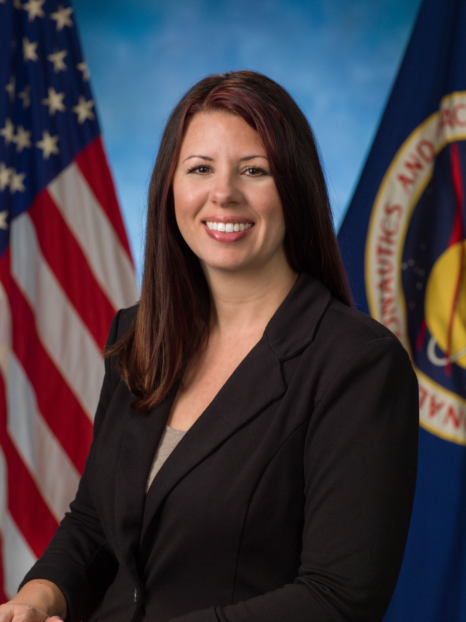 A woman dressed in a black suit stands in front of a blue background.