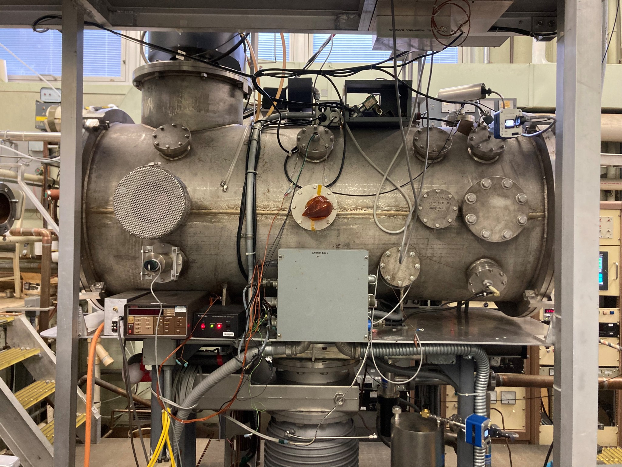 A large cylindrical vacuum chamber with pipes, wires, and control panels in a metal frame, surrounded by instruments.