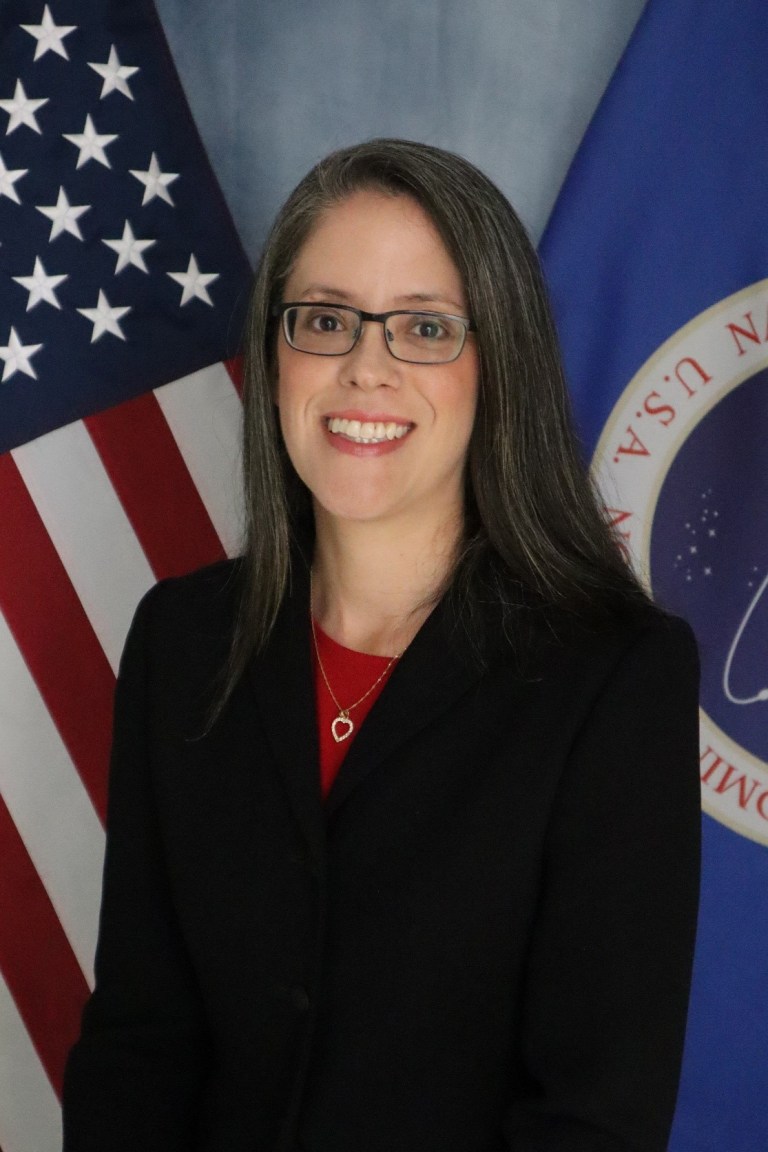 Image shows woman in black suit with red top posing for a photo with an American flag and NASA flag.