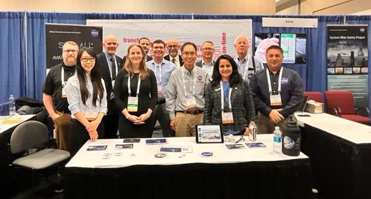 Image of 12 smiling people standing behind a table with NASA swag, and behind them are some posters explaining some of the things they are working on, with the caption, "DIP TEAM at ATCA Global Conference"