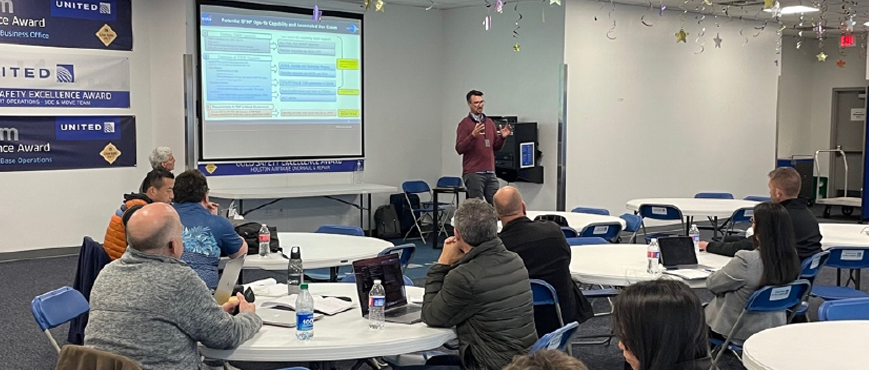 Image of 9 people sitting, and 1 giving a presentation on airspace management technology use cases, with the caption "Jeremy Coupe out-briefing stakeholders in Houston"