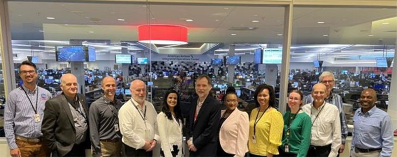 Image of 12 smiling people standing faced towards the camera, and behind them is a big room with many monitors with a sign saying "American Airlines Integrated Operations Center", and with the caption "FAA and NASA at American Airlines IOC"