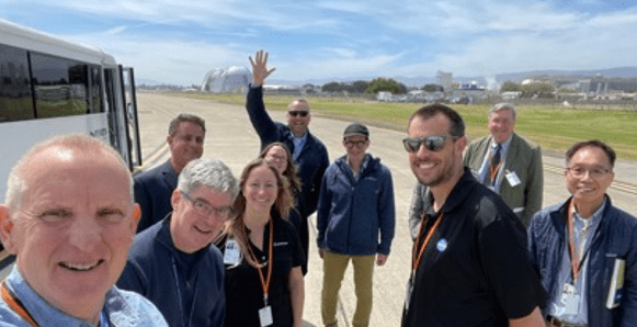 Image of 10 people smiling, and one waving, outside on a sunny day with an airfield in the background, with the caption, "Site Visit at Moffett Field Airport"