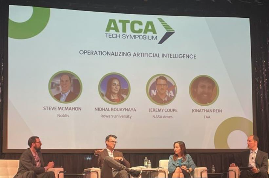 Image of 4 people sitting and discussing something, and in the background is a large screen reading "ATCA: Tech Symposium: Operationalizing Artificial Intelligence", with the caption "Operationalizing AI Panel at ATCA 2024. Dr Coupe is 2nd from left"