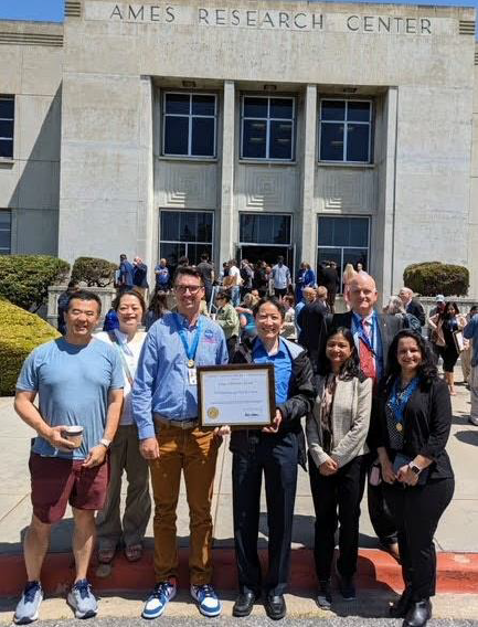 Image of 7 people smiling in front of a building named "Ames Research Center", and they are holding an award, with the caption "From left to right: Liang Chen, Miwa Hayashi, Jeremy Coupe, Gilbert Wu, Swati Saxena"