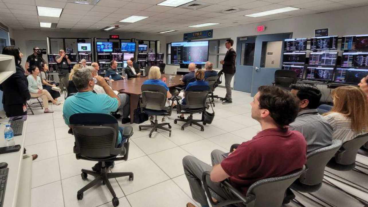 Image of a room full of people looking at a person standing and giving a presentation in a room full of screens with air traffic information on them, with the caption "Dr. Jeremy Coupe briefed the NAC Aeronautics Committee about the SFNP-Ops demonstration"