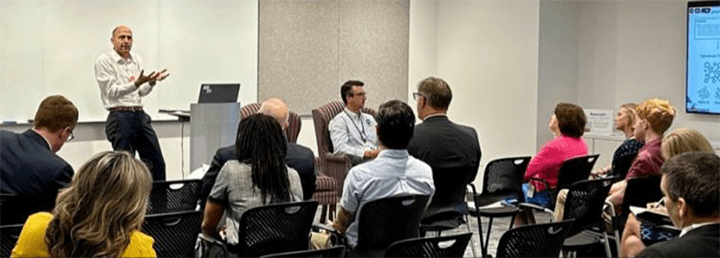 Image of a man giving a presentation in front of a room of people, with the caption "Dr. Tim Niznik of American Airlines (left on the stage) and Dr. Jeremy Coupe of NASA DIP (right, seated, on the stage)