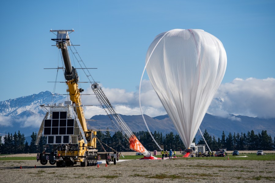 A large metal scientific instrument with black panels is suspended off the ground by a yellow crane. A white trail of material is on the ground and attaches to a partially inflated white balloon in the background.