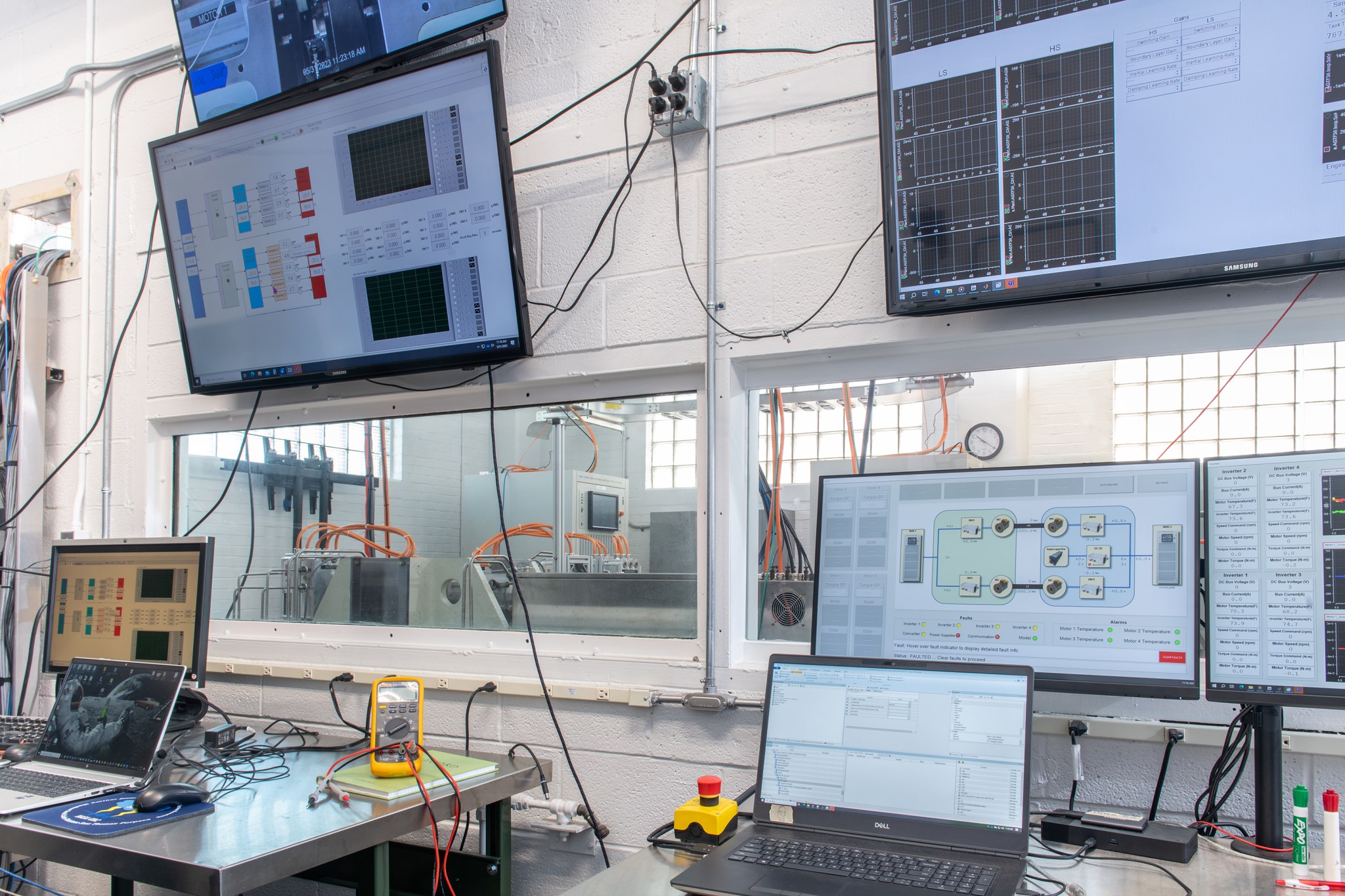 A set of laptops placed on a desk with monitors attached to the wall above overlook a testing rig through a clear window.