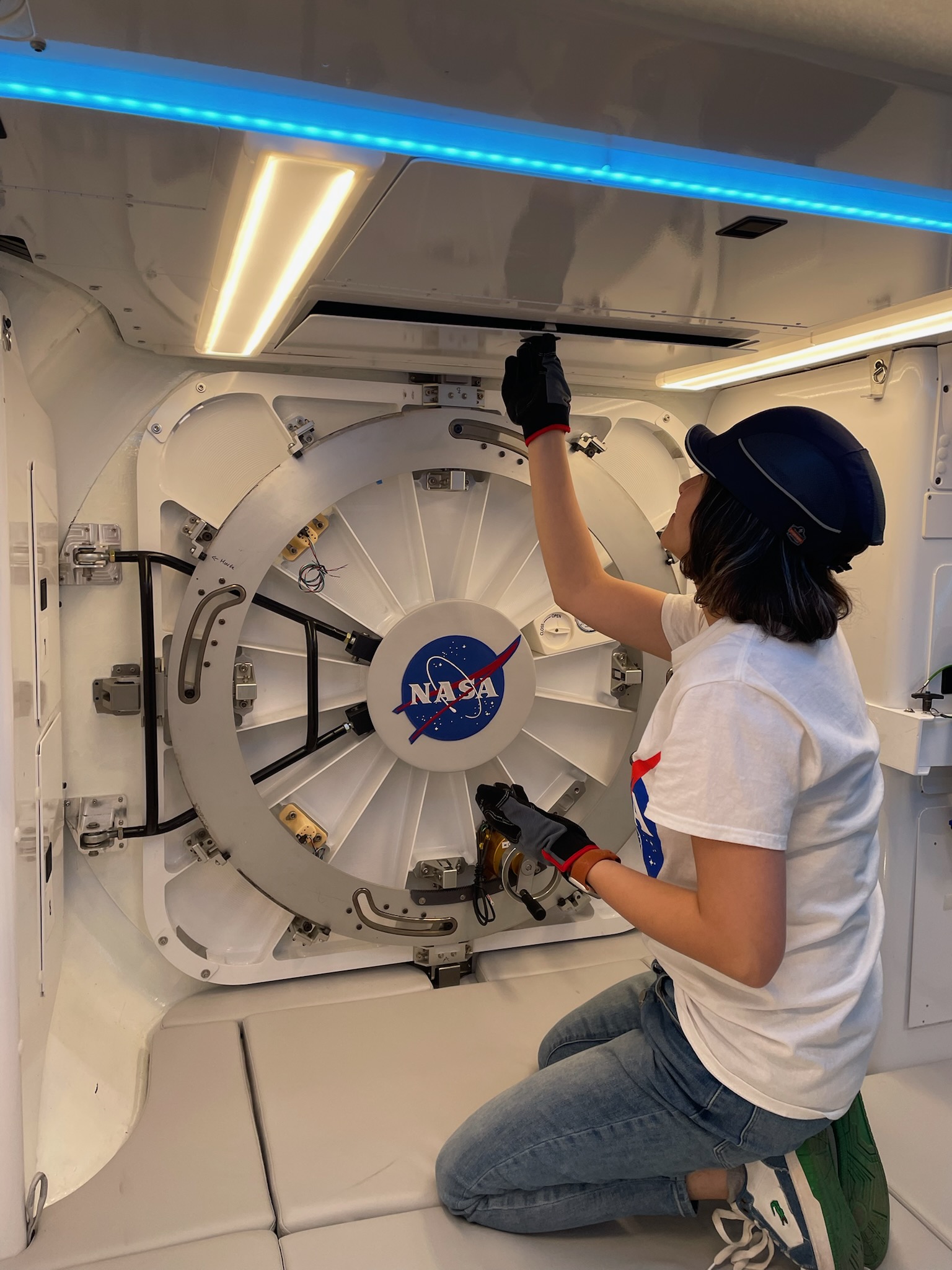 A woman wearing casual clothes and work gloves screws a metal panel down inside of a space vehicle mockup.