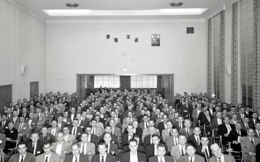 People seated in auditorium and foyer in background.