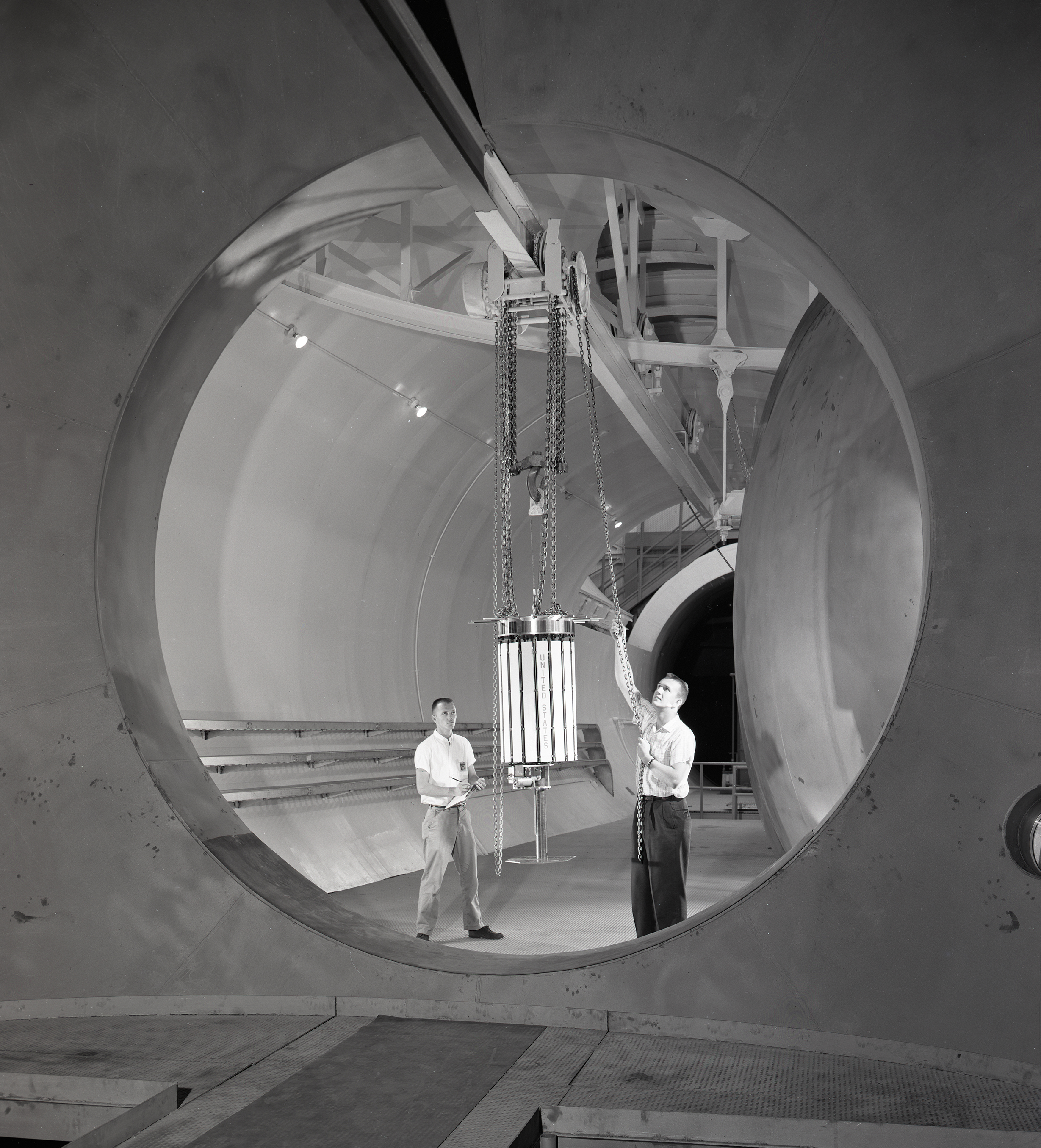 Two men move satellite with crane in chamber.